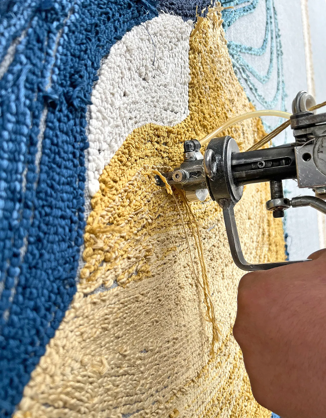 Hand using tufting gun to create a textured textile art piece with blue, white, and beige yarn.