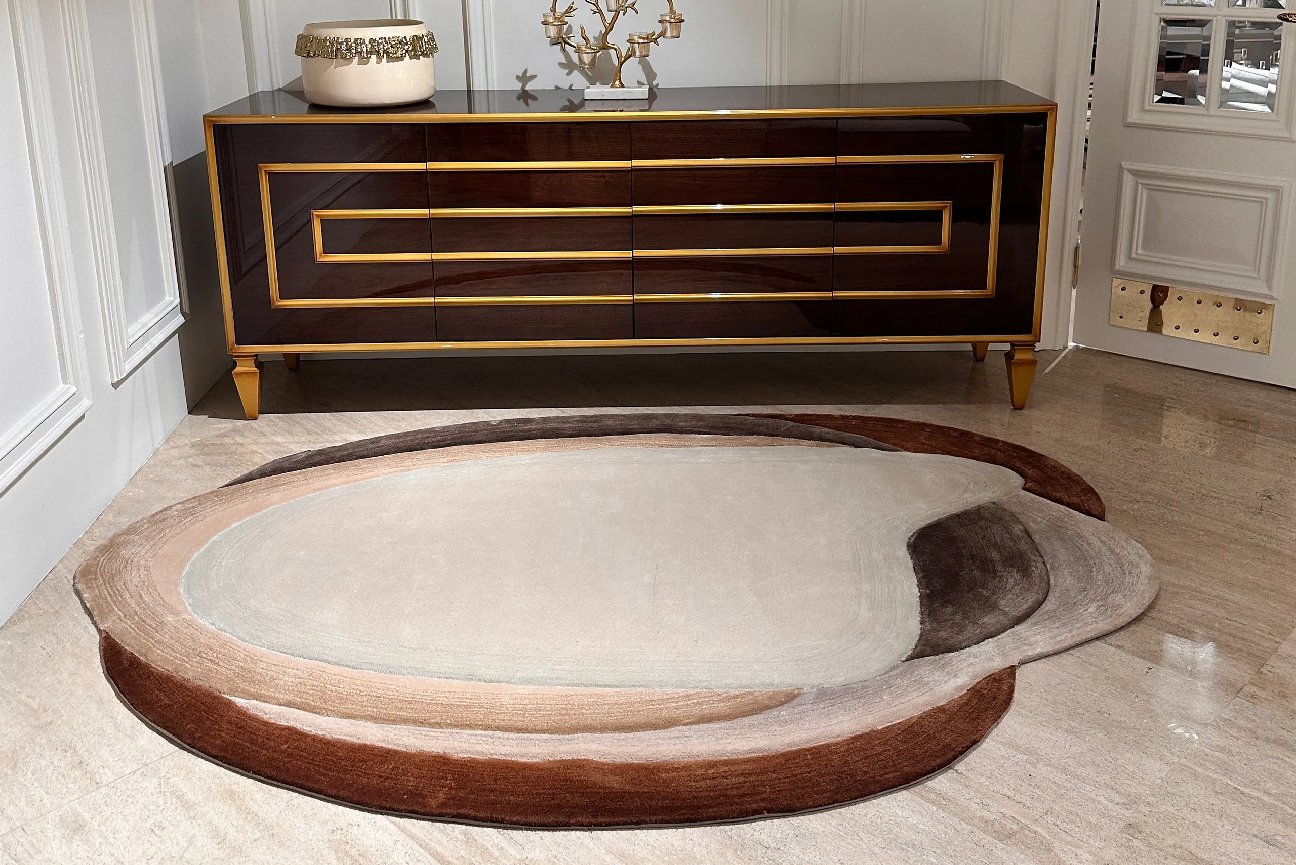 Oval area rug with layered shades of beige, brown, and cream on a polished floor in front of a dark wood sideboard with gold trim.