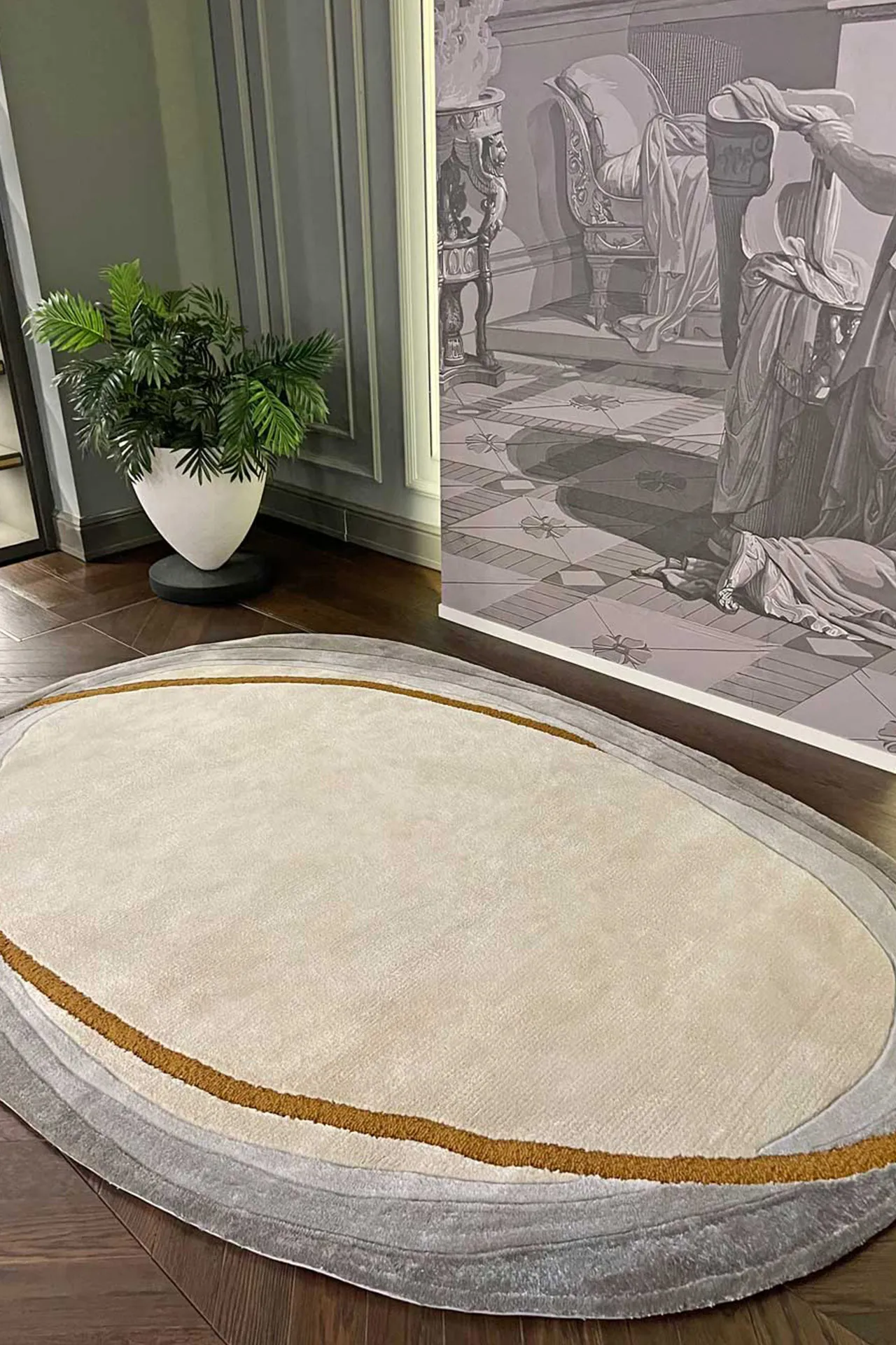 Oval beige rug with gray and brown layered border on wooden floor, next to a leafy green plant in a white pot and monochrome wall art.