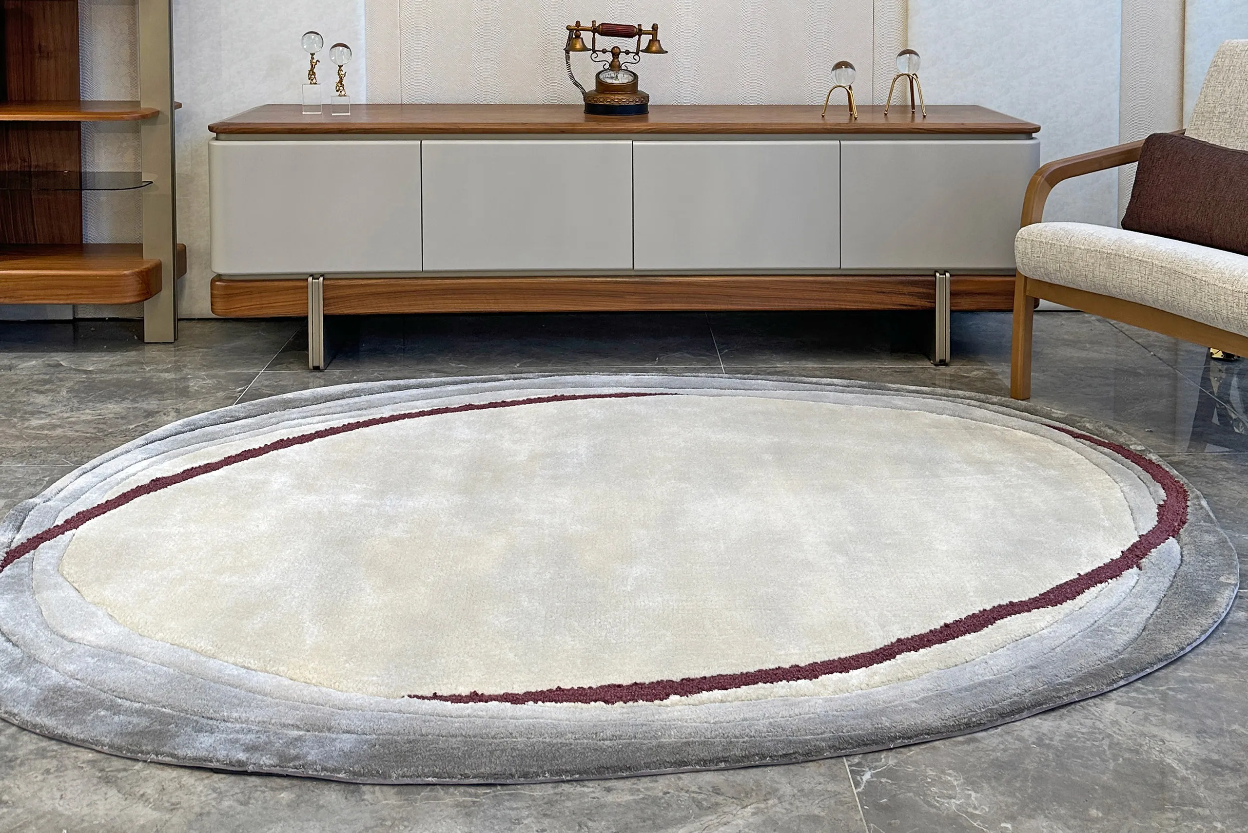 Oval beige and gray area rug with a curved maroon line in a modern living room with a light gray cabinet and a beige armchair.